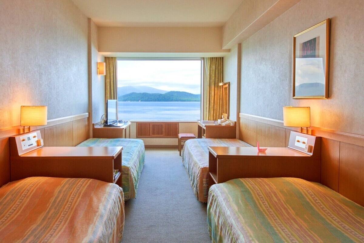 A hotel room with two beds, a desk, a chair, a window with a view of the sea, and a framed picture on the wall.