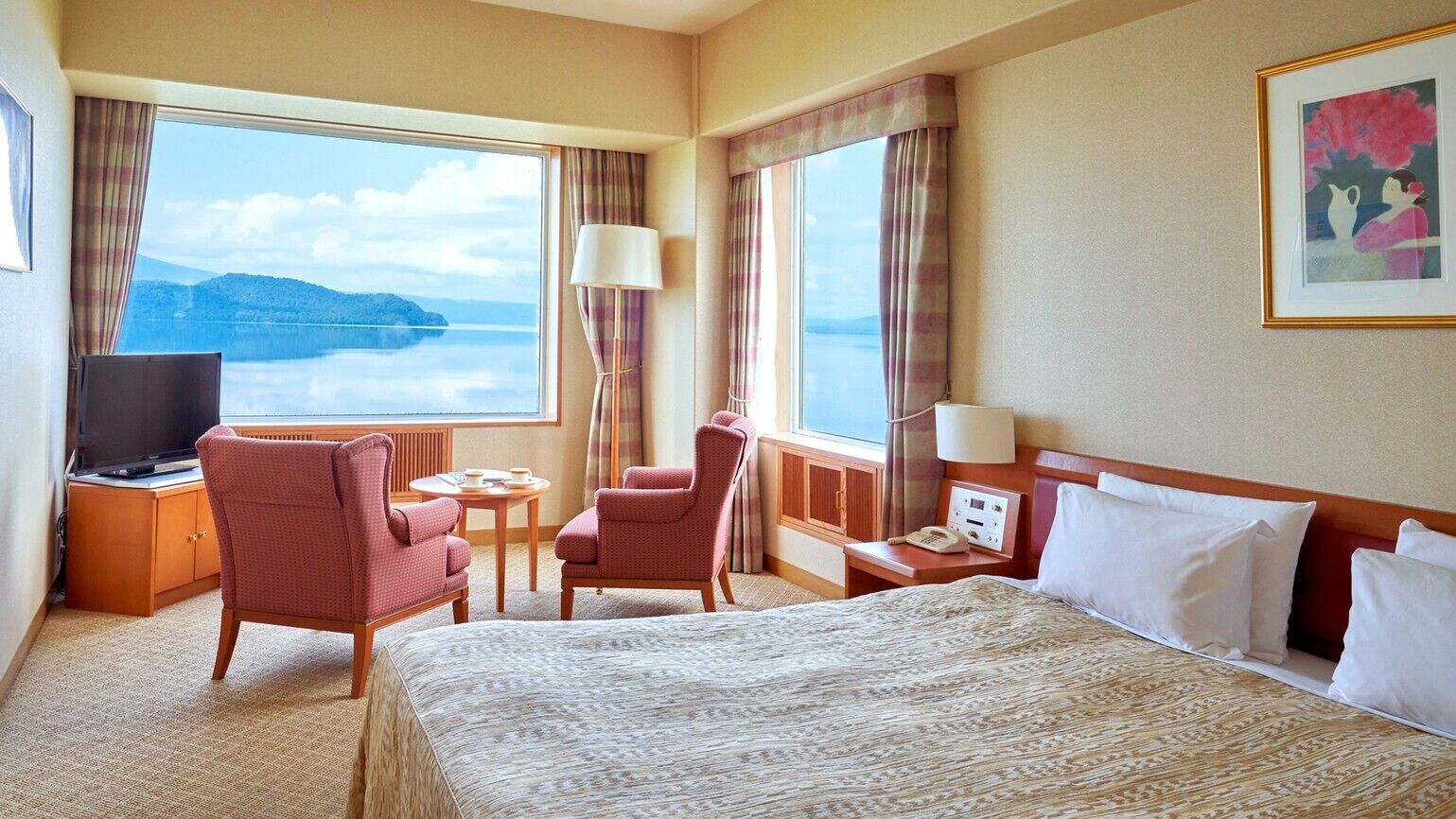 A hotel room with a bed, a TV, a desk with a lamp, a chair, and a view of the water.