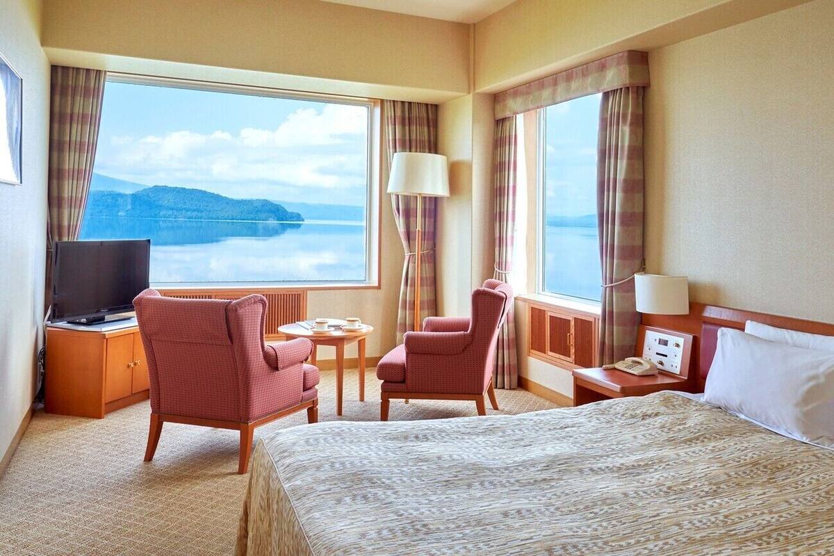A hotel room with a bed, a TV, a desk with a lamp, a chair, and a view of the water.