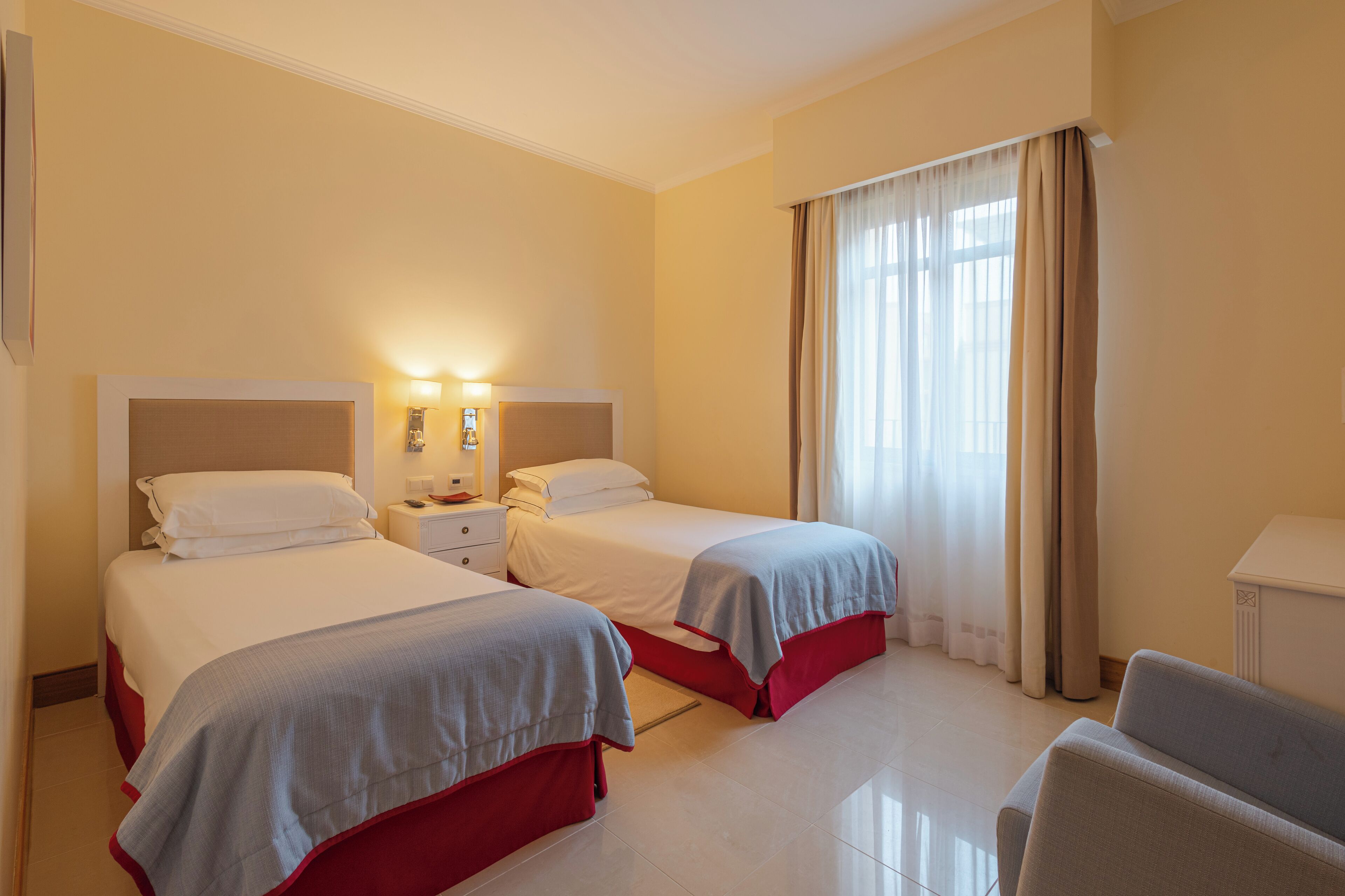 Una habitación de hotel con dos camas, una mesita de noche, una lámpara, una silla y una ventana con cortinas.