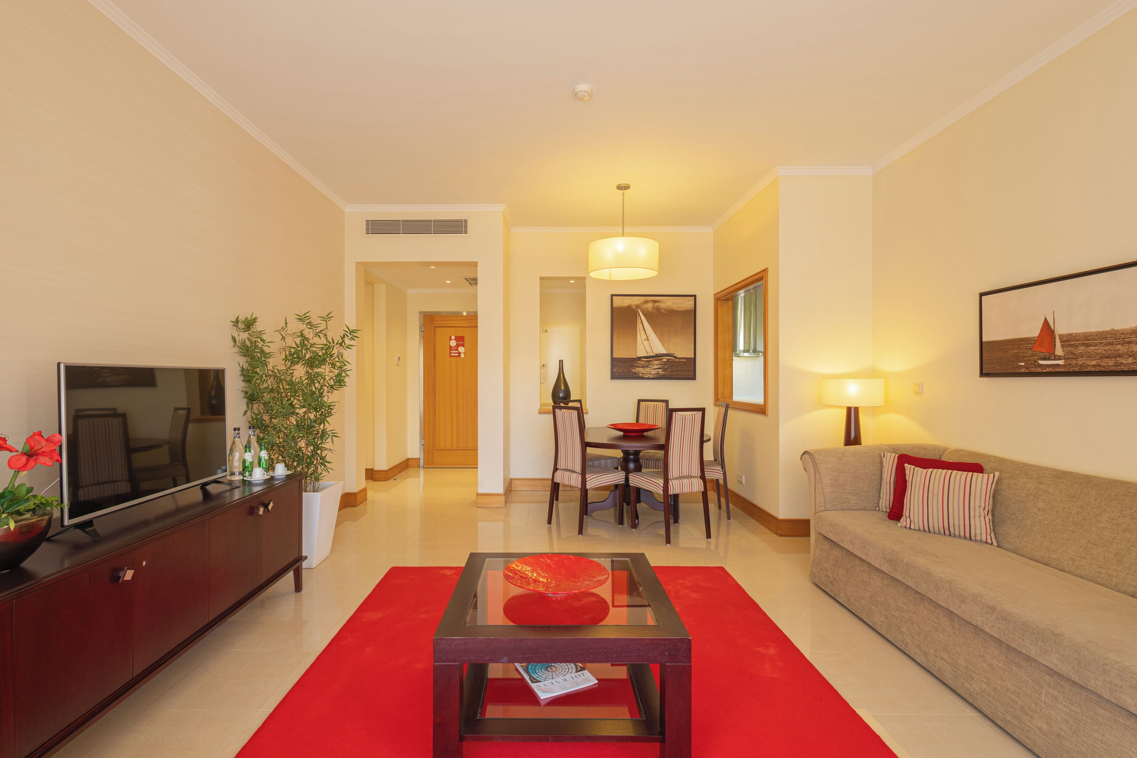 A modern living room with a flat-screen TV, a red sofa, a glass coffee table, and a dining area with a chandelier.