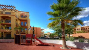 A playground with a slide and climbing structures, a palm tree, and a building with balconies and arched windows. - The Residences Vilamoura (Loulé)