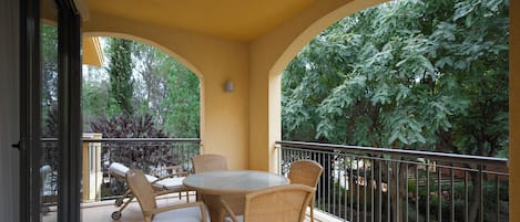 A balcony with wicker furniture, a glass table, and a view of trees.