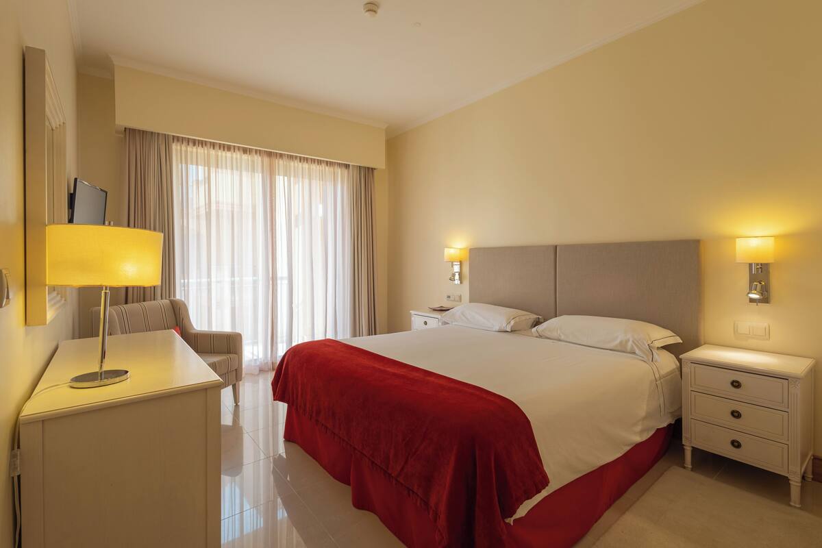 Ein Hotelzimmer mit einem großen Bett, einem Schreibtisch mit Lampe, einem Sessel, einem Nachttisch mit Schubladen und einem Fenster mit Vorhängen.
