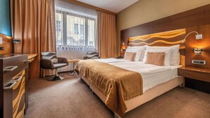 A hotel room with a large bed, a desk, a chair, and a view of the city through the window. - Hotel Danubia Gate (Bratislava)