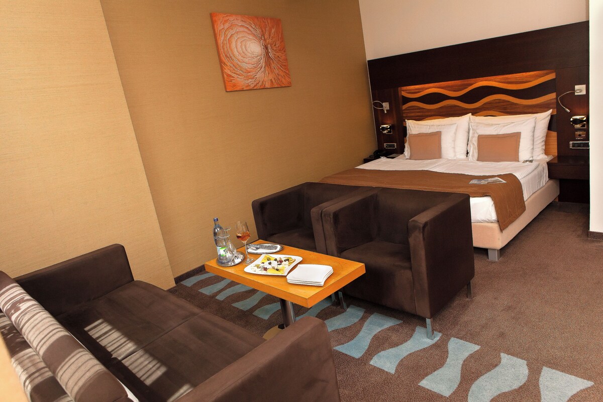 A hotel room with a bed, two armchairs, a small table with a bottle, glasses, and a plate of food.