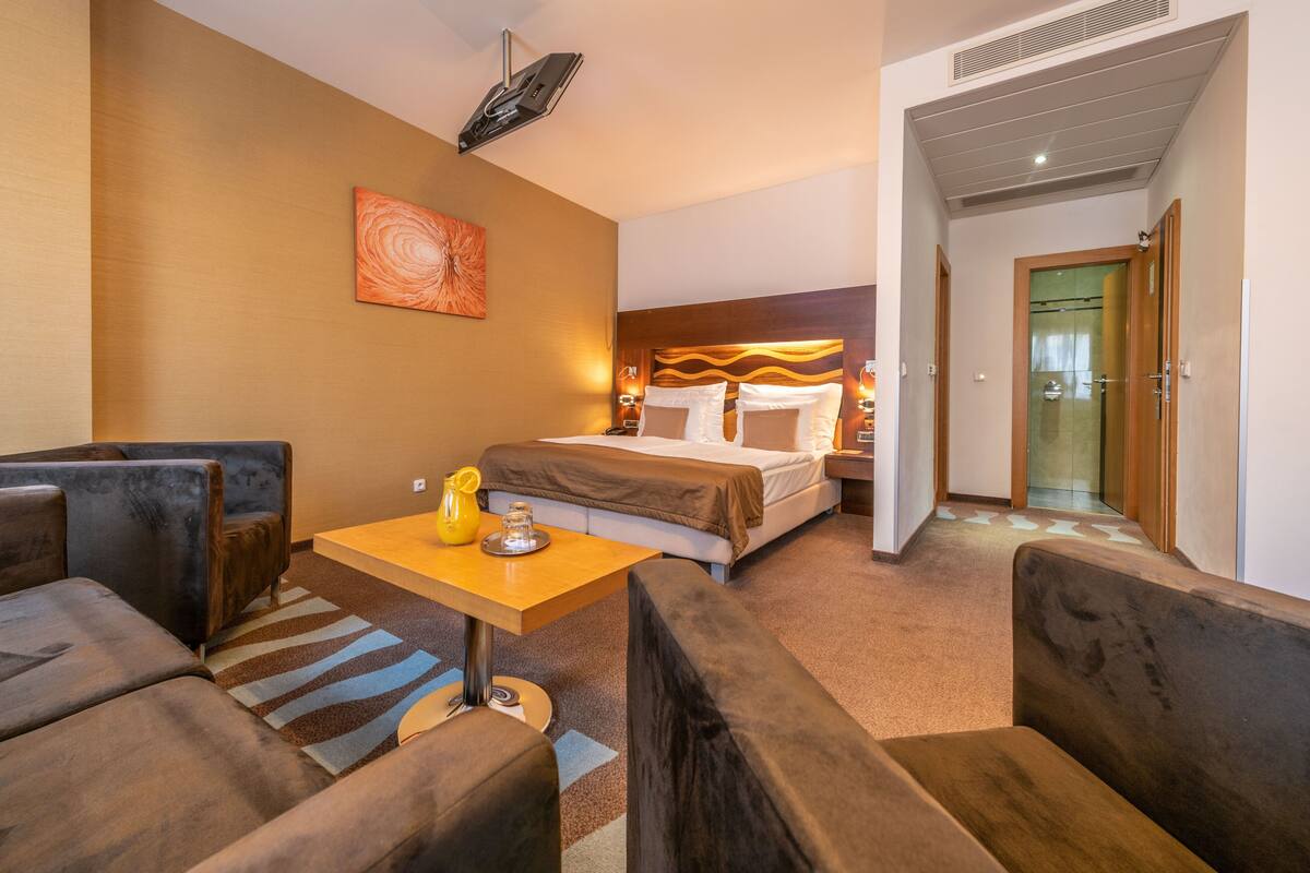 A hotel room with a bed, a sofa, a small table with a lamp, and a painting on the wall.