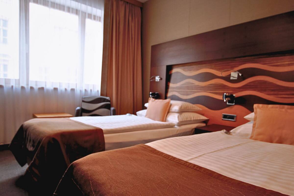 A hotel room with two beds, a large window with curtains, a headboard with a wavy design, and a chair.