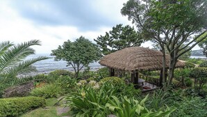 Un pavillon au toit de chaume, doté d’un banc, entouré d’une végétation luxuriante et offrant une vue sur une étendue d’eau.