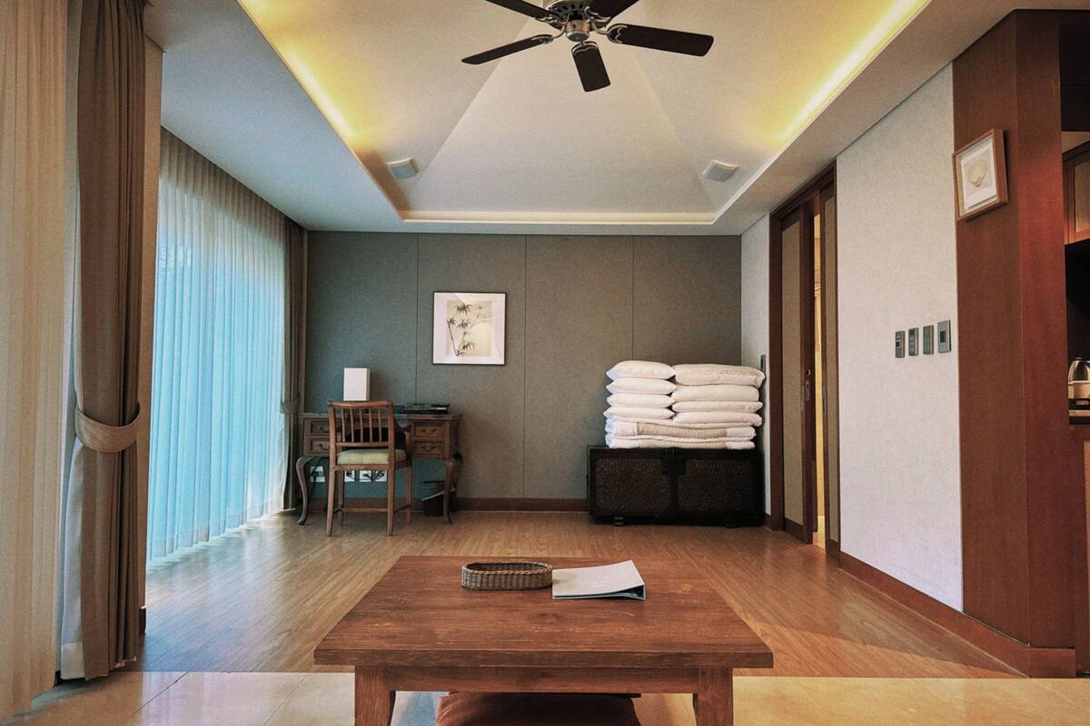 Une chambre d’hôtel avec un ventilateur de plafond, une table en bois sur laquelle se trouvent un panier et un livre, un bureau avec une chaise, une pile de serviettes et un tableau encadré au mur.