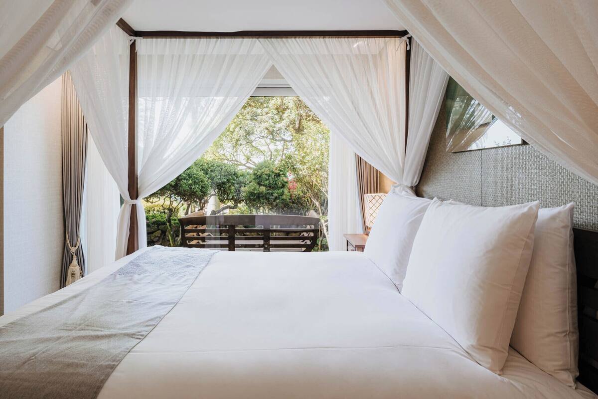 Une chambre à coucher avec un lit à baldaquin, une vue sur la verdure et un banc en bois.