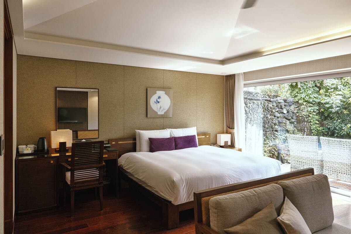 Une chambre d’hôtel équipée d’un lit, d’un bureau, d’une chaise et d’une grande fenêtre donnant sur un paysage verdoyant.