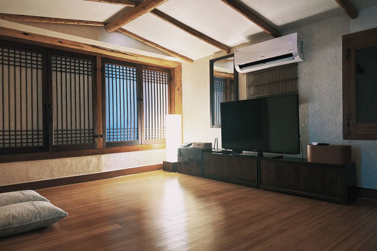 Une pièce avec un plancher en bois, un téléviseur posé sur un meuble, une grande fenêtre donnant sur un beau paysage et un ventilateur de plafond.