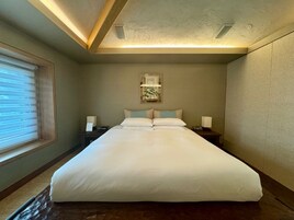 A neatly made bed in a hotel room with a large window, a bedside table with a lamp, and a framed artwork on the wall.