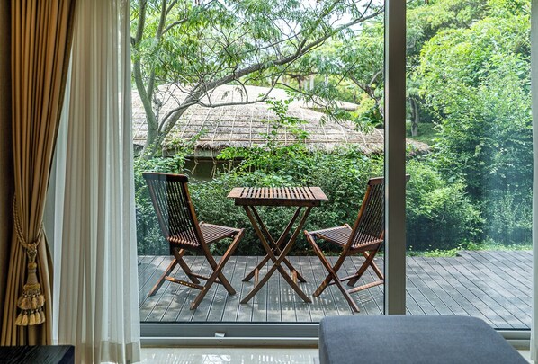 Un balcon avec deux chaises en bois et une table, donnant sur un jardin luxuriant et une construction à toiture de chaume.