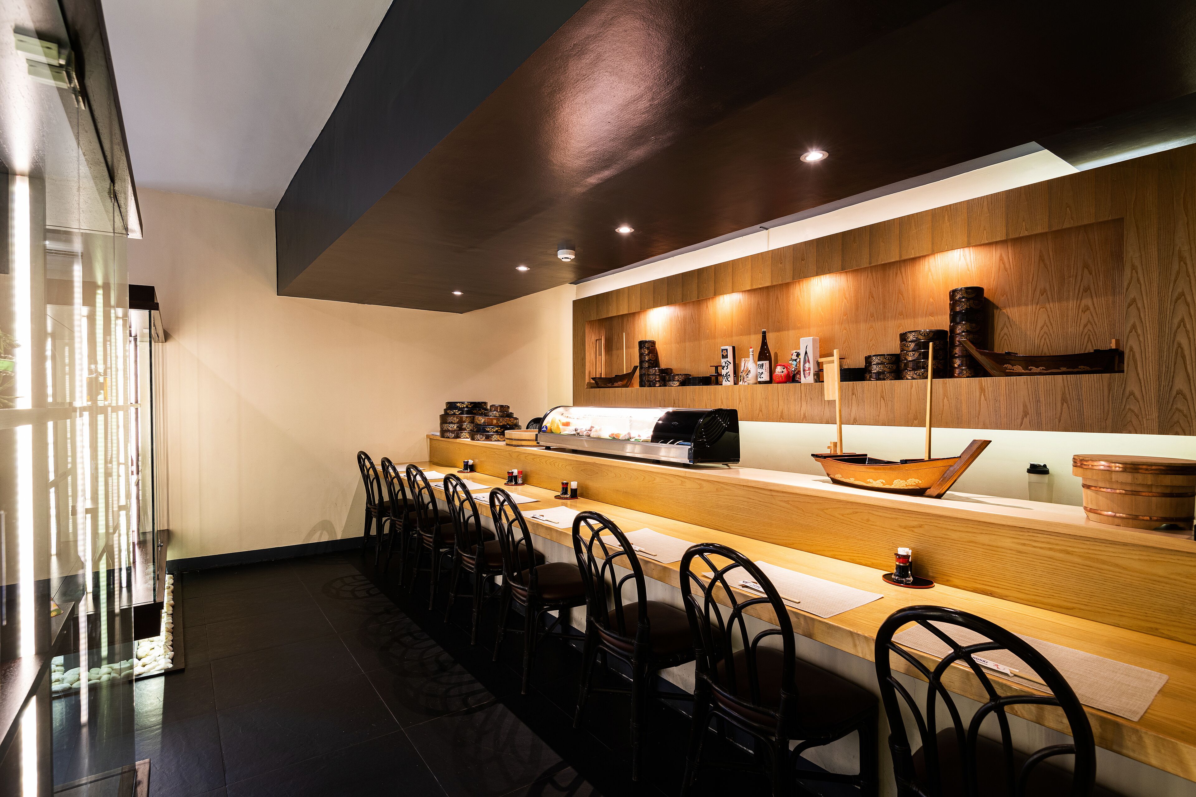 A modern bar area with wooden shelves, a long counter, and several black chairs.