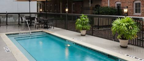 A swimming pool with a brick building in the background, outdoor seating with umbrellas, and potted plants.