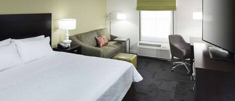 A hotel room with a large bed, a desk with a flat-screen TV, a chair, a lamp, and a window with a green blind.