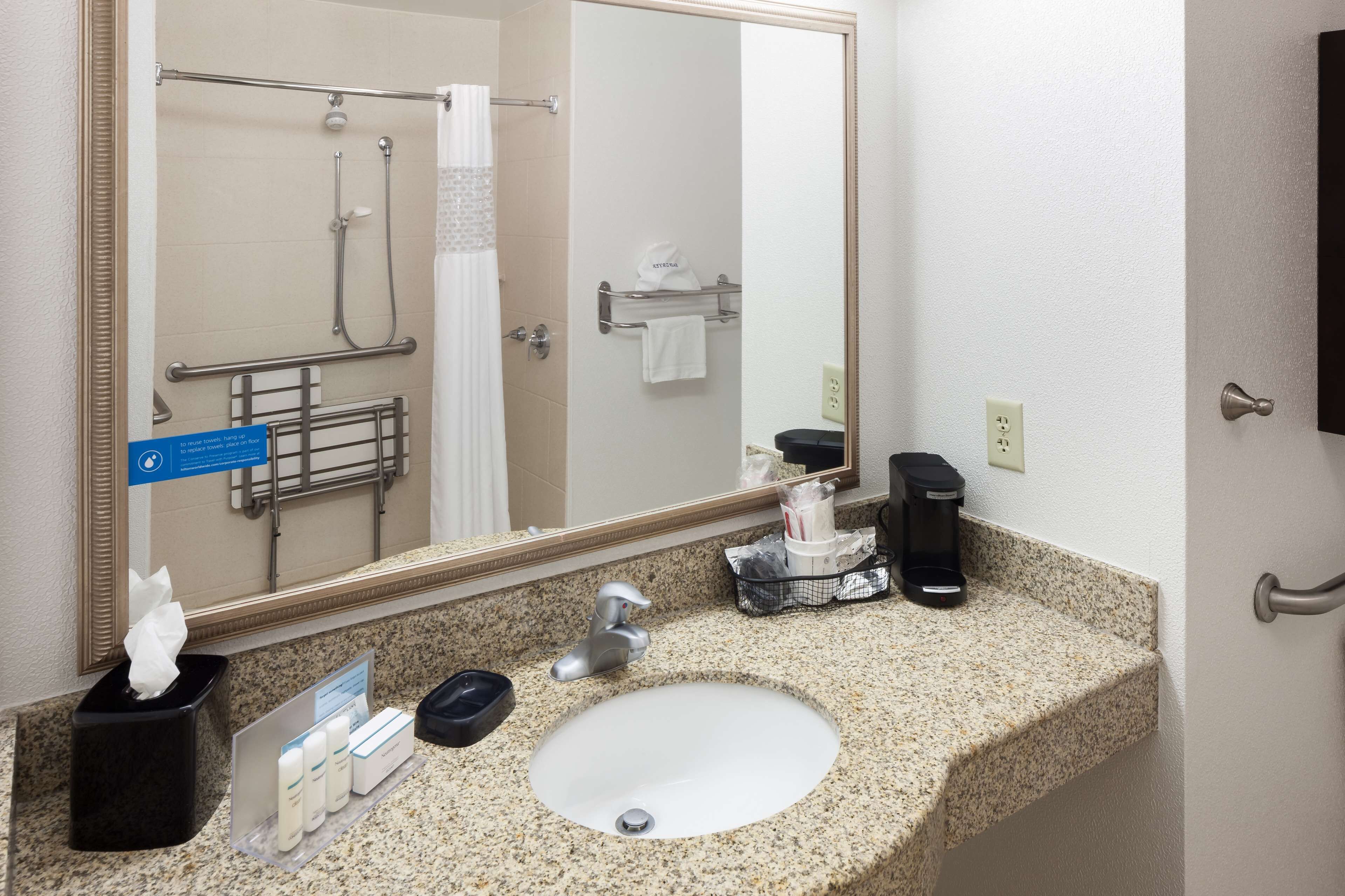 Un baño de hotel con lavamanos de encimera de granito, un espejo grande, toallero, zona de ducha y cafetera.