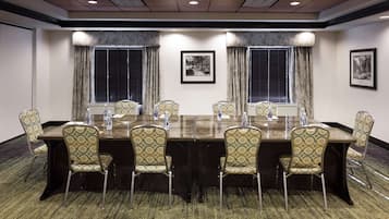 A conference room with a long table, six chairs, two windows with blinds, and framed pictures on the walls.