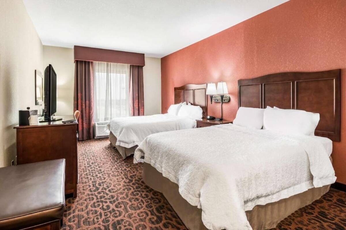 Ein Hotelzimmer mit zwei Betten, einem Schreibtisch, einem Flachbildfernseher, einem Sessel und einem gemusterten Teppich.