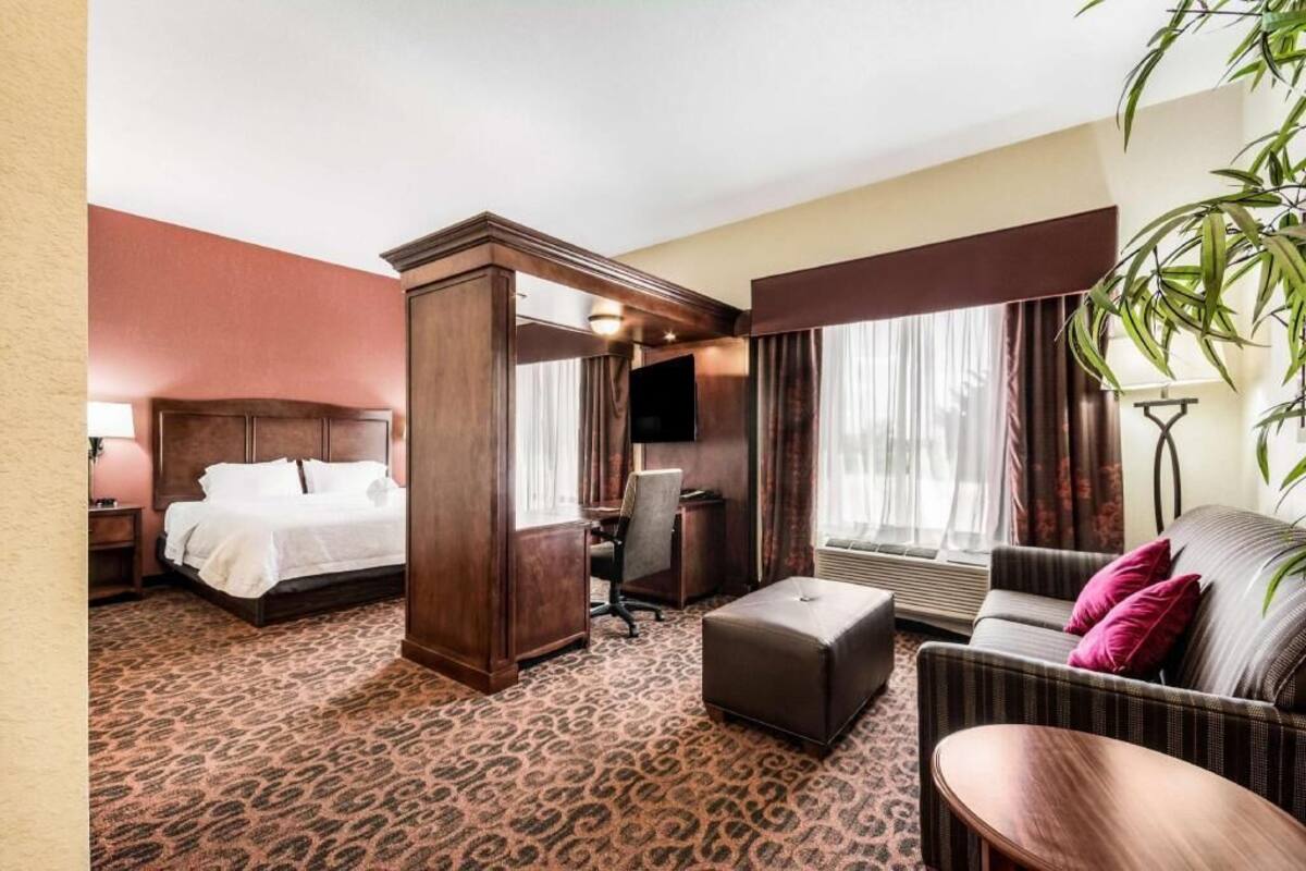 Ein Hotelzimmer mit einem Bett, einem Schreibtisch, einem Sofa, einem Hocker und einem großen Fenster mit Vorhängen.