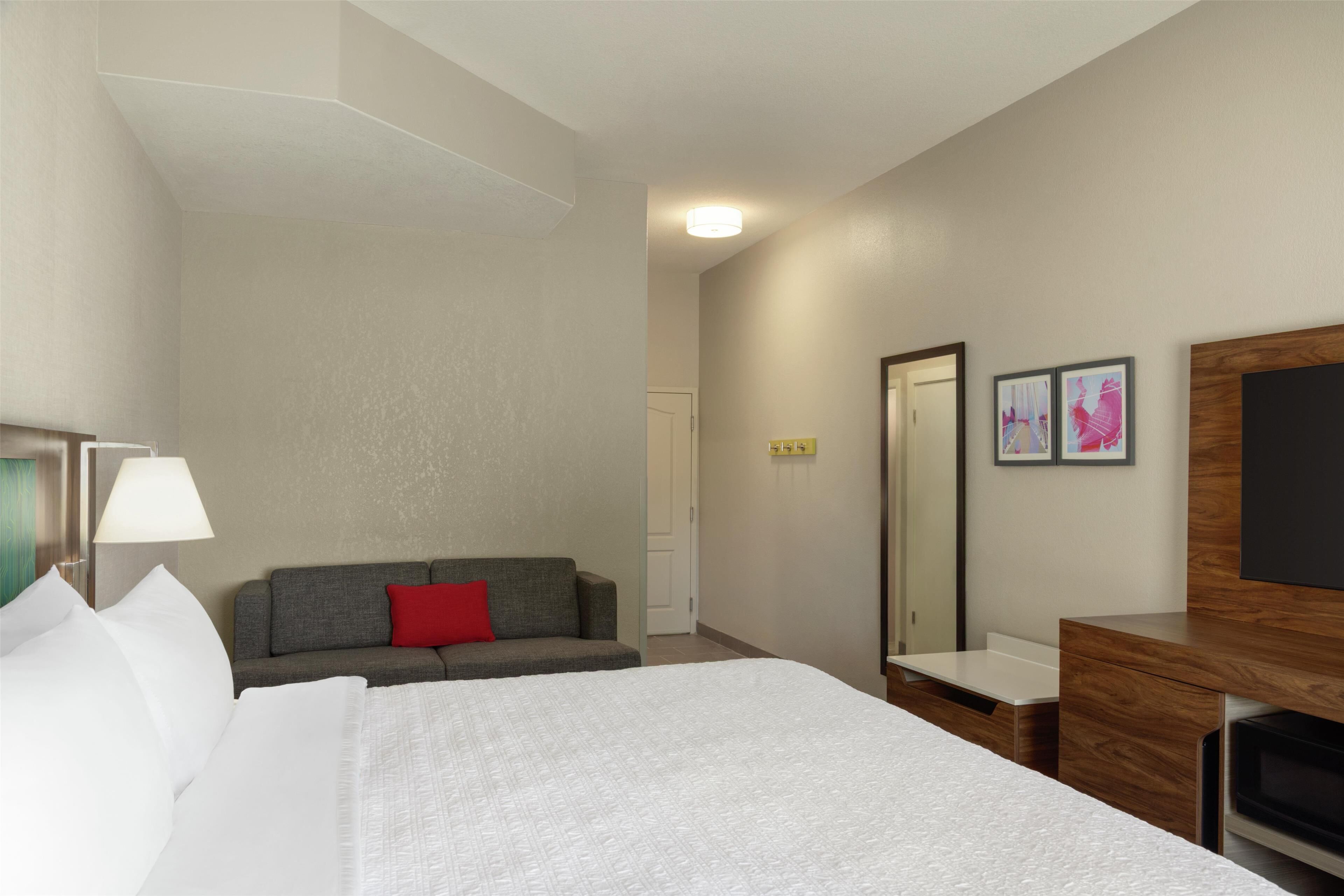 Habitación de hotel con una cama, un sofá gris con almohada roja, un aparador de madera, un espejo y dos cuadros en la pared.