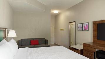 Habitación de hotel con una cama, un sofá gris con almohada roja, un aparador de madera, un espejo y dos cuadros en la pared.
