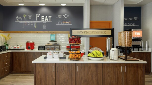 A breakfast bar with a sign reading "HAPPY BREAKFAST STARTS HERE" and various food items like fruits and pastries.