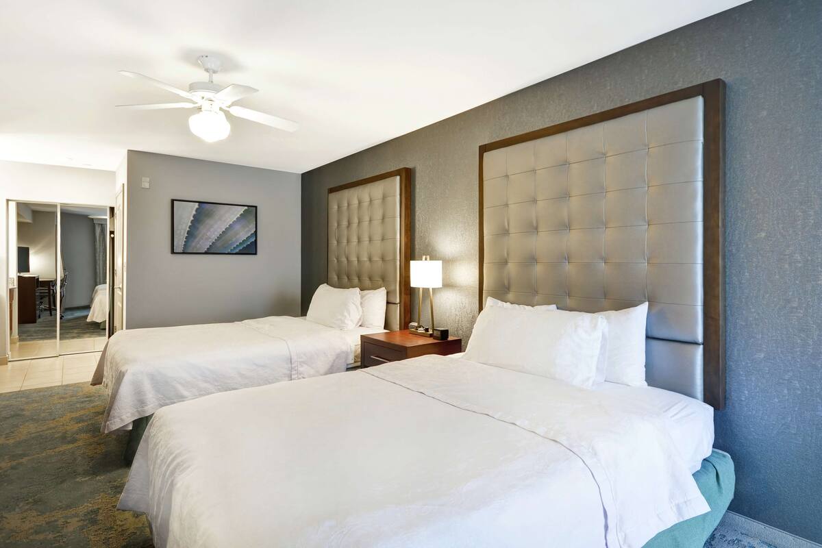 Een hotelkamer met twee bedden, een plafondventilator, een nachtkastje met een lamp en een ingelijste foto aan de muur.