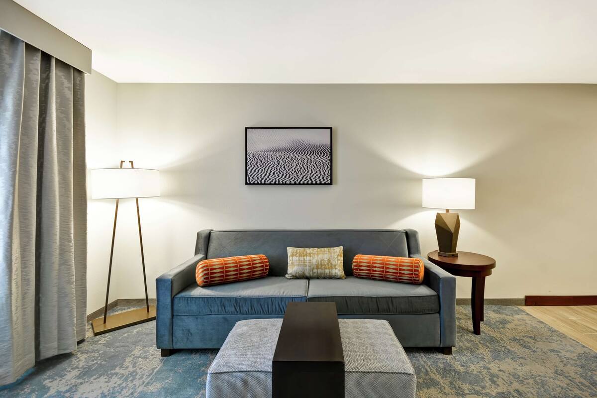 Een moderne hotelkamer met een blauwe bank, twee bijzettafels met lampen, een lijstwerk abstract kunstwerk en een kamerbreed tapijt met dessin.
