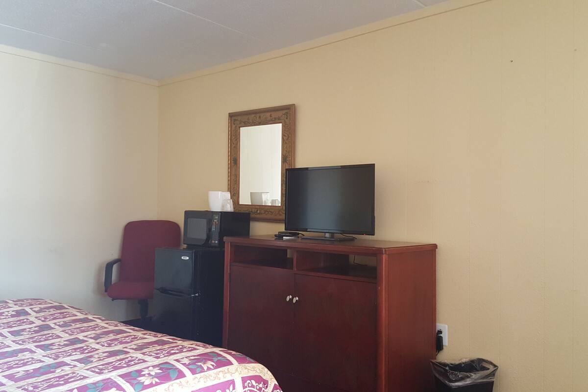 A hotel room with a bed, a TV on a wooden cabinet, a red chair, a microwave, and a trash bin.