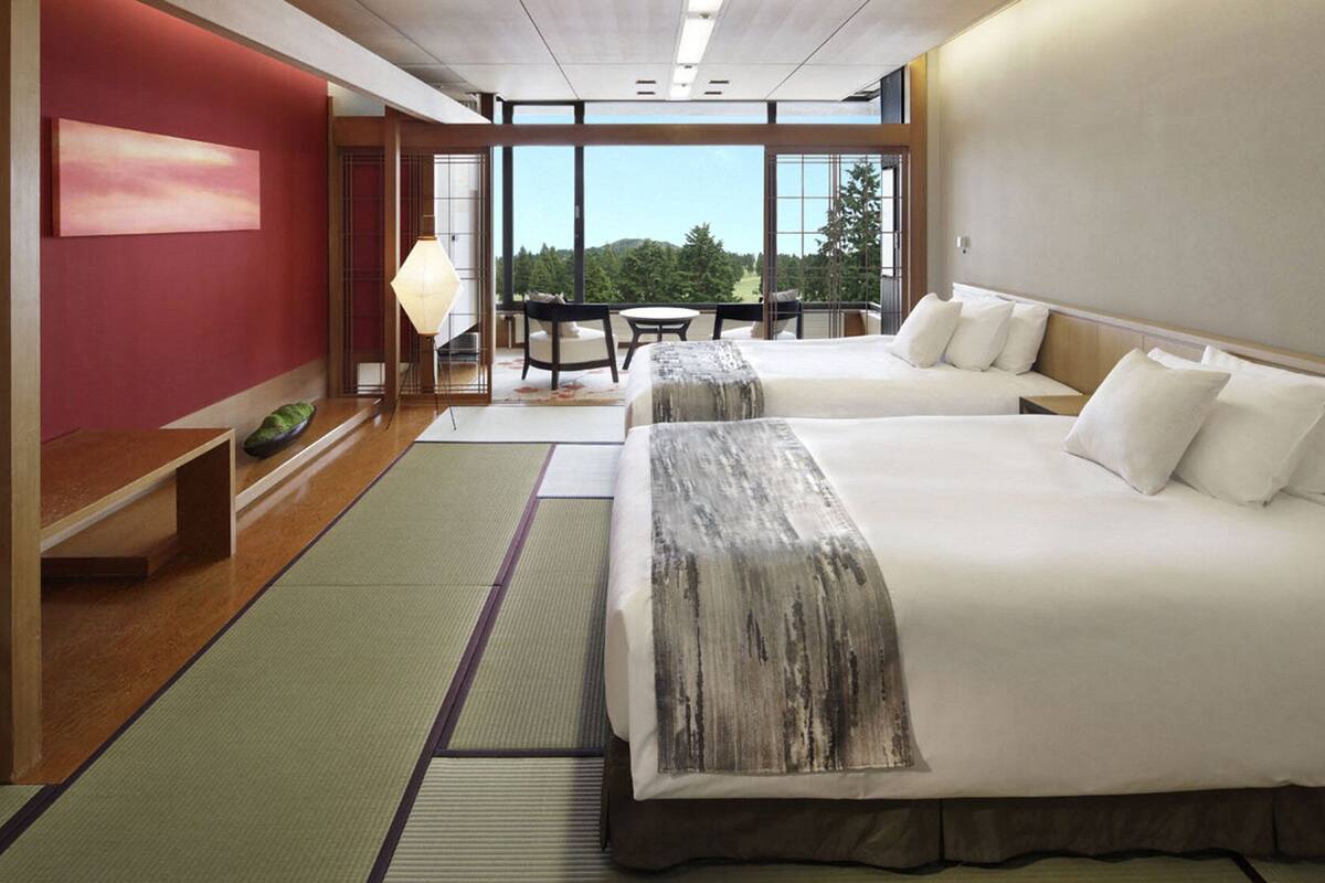 A hotel room with two beds, a view of trees, a dining area, and a red wall.