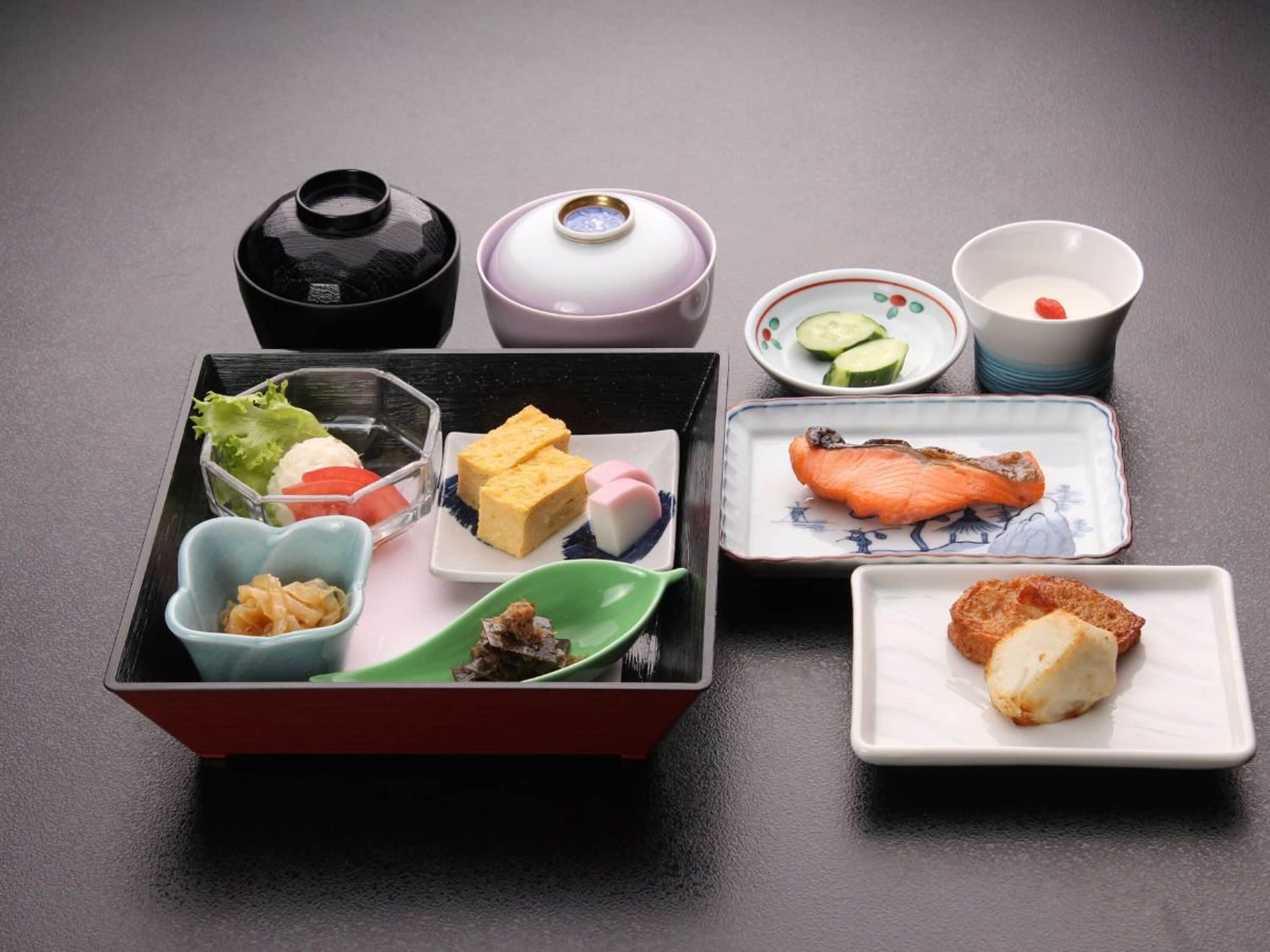 Una caja bento japonesa tradicional con diversos alimentos, como pescado, verduras y arroz, acompañada de pequeños platos y un juego de té.