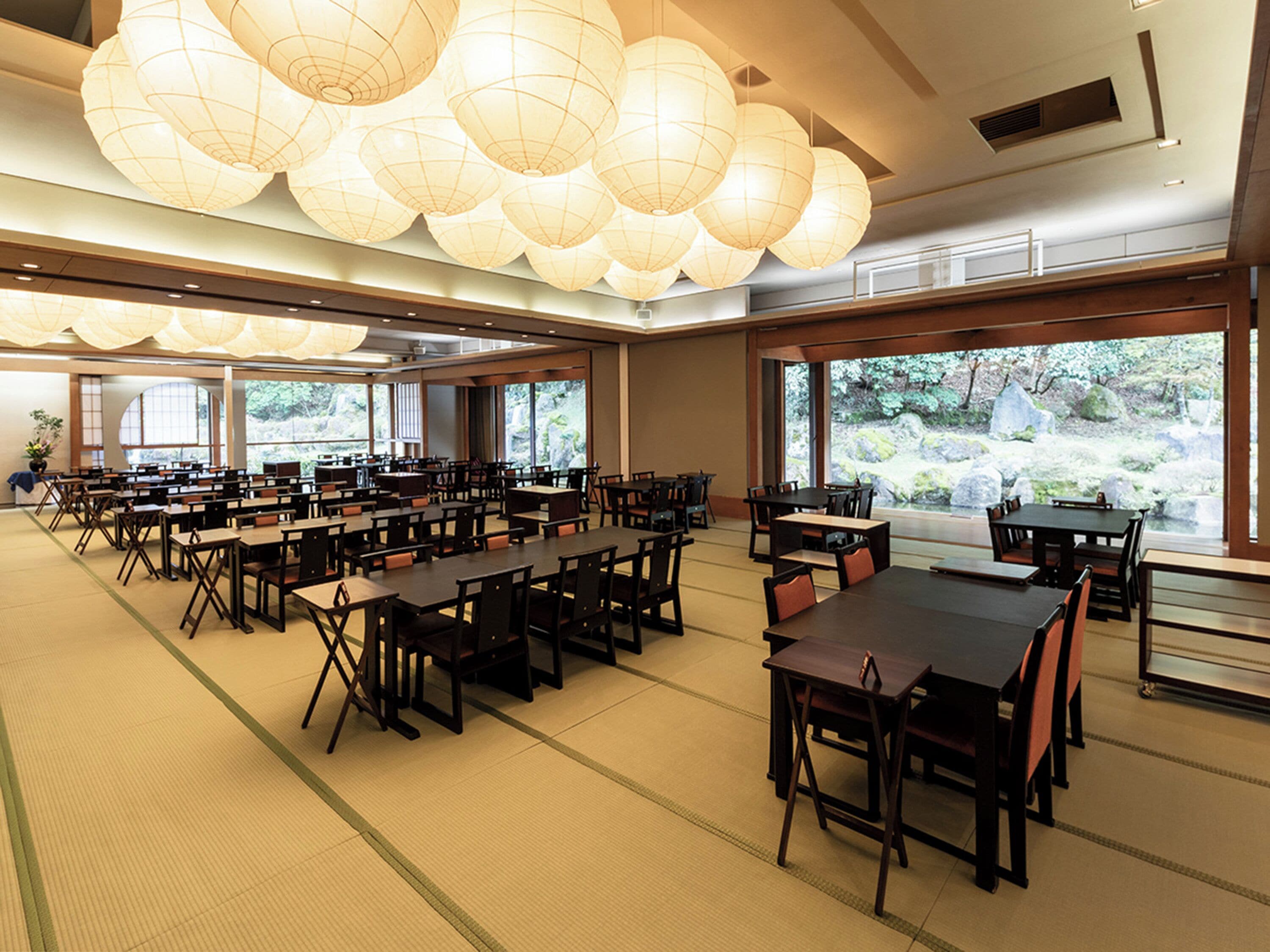 A spacious dining area with multiple tables and chairs, large windows offering a view of a natural landscape, and traditional paper lanterns hanging from the ceiling.