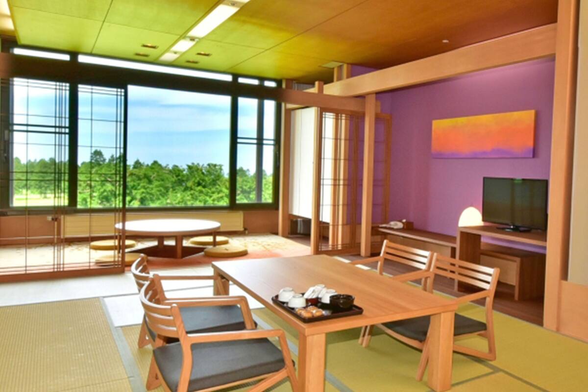A modern dining area with a wooden table and chairs, a television, and a view of greenery through large windows.