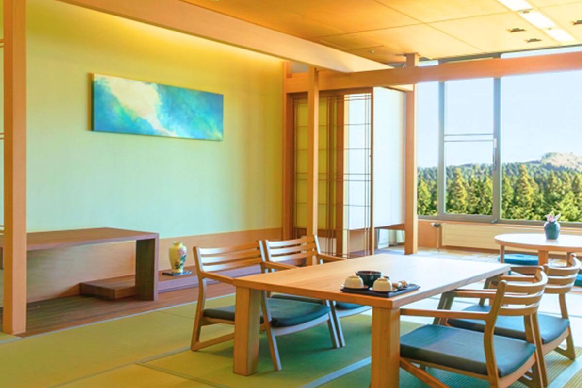 A dining area with a wooden table and chairs, a painting on the wall, and a view of a forest through large windows.
