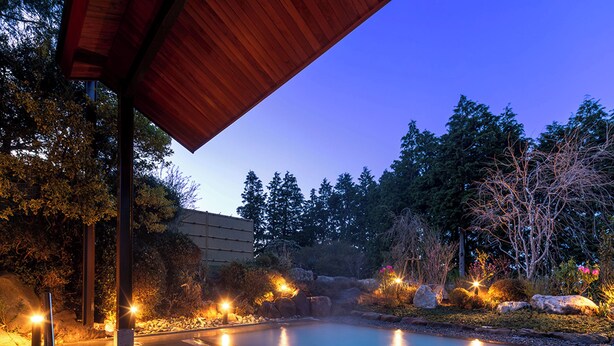 Un área de piscina exterior tranquila con un porche de madera, rodeada de abundante vegetación e iluminada con luces ambientales.
