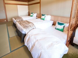 Una habitación de hotel con tres camas, cada una con ropa de cama blanca y almohadas verdes y blancas. La habitación presenta una estética japonesa tradicional con suelo de tatami y cabeceros de madera.
