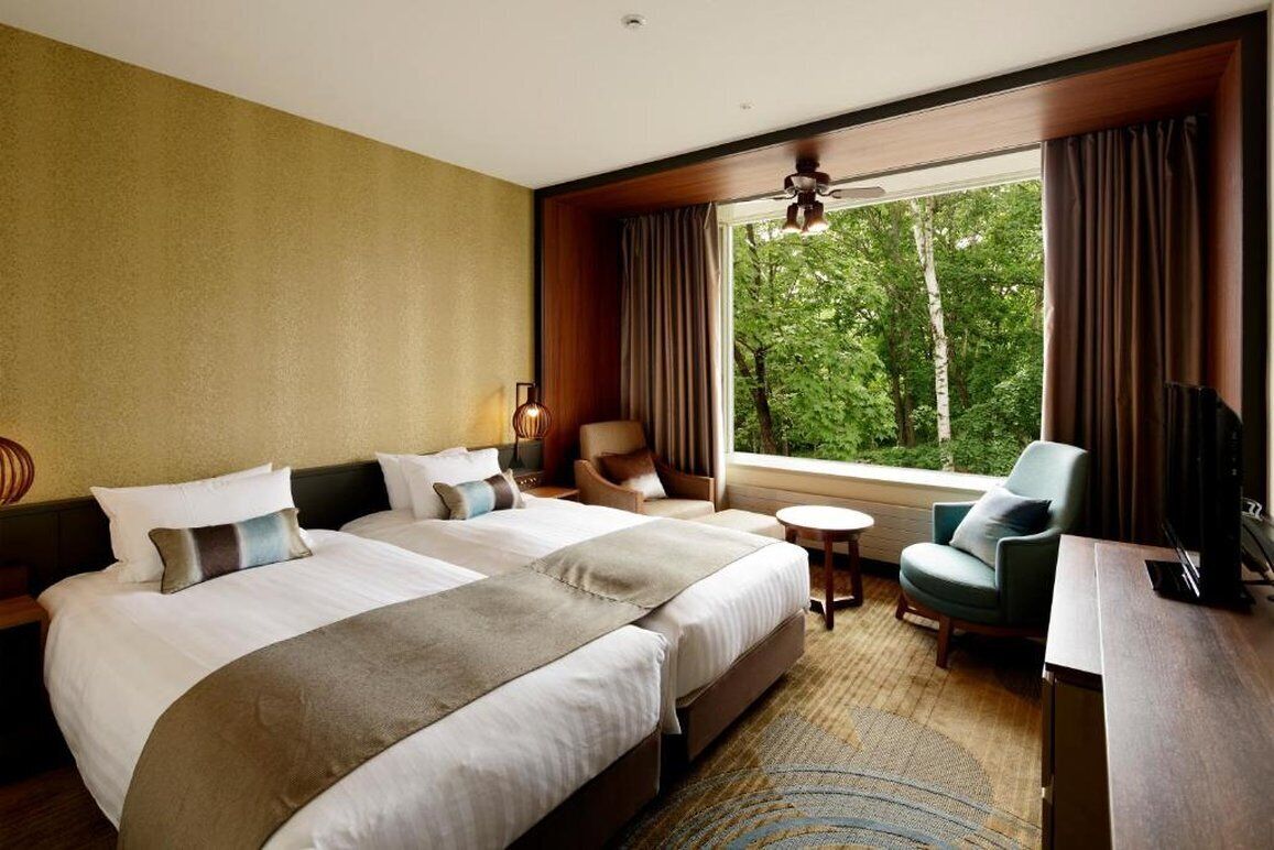 Une chambre d’hôtel avec deux lits, un canapé, une télévision et une fenêtre donnant sur des arbres.