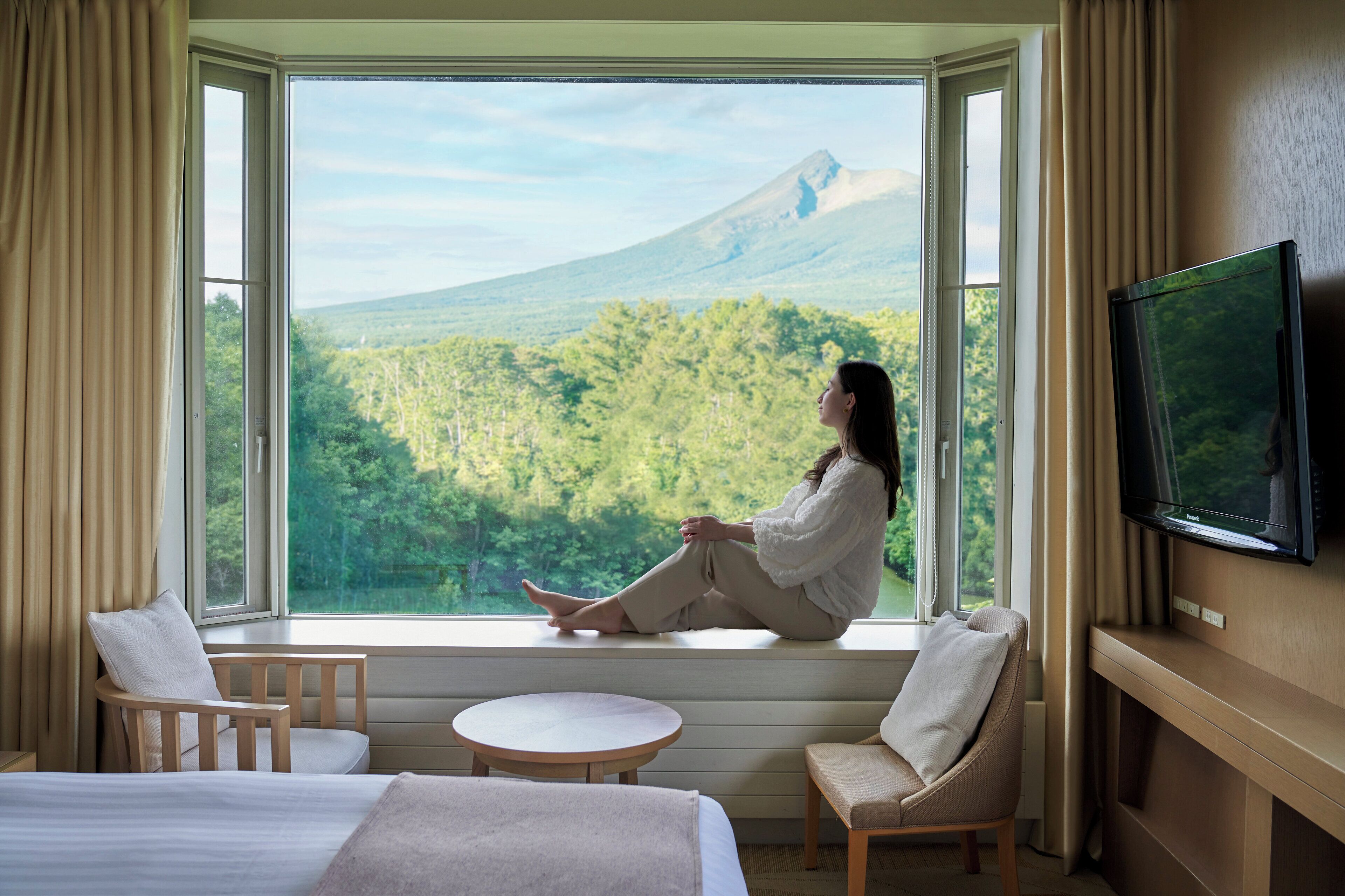 A hotel room with a large window, a bed, two chairs, a small table, and a TV.