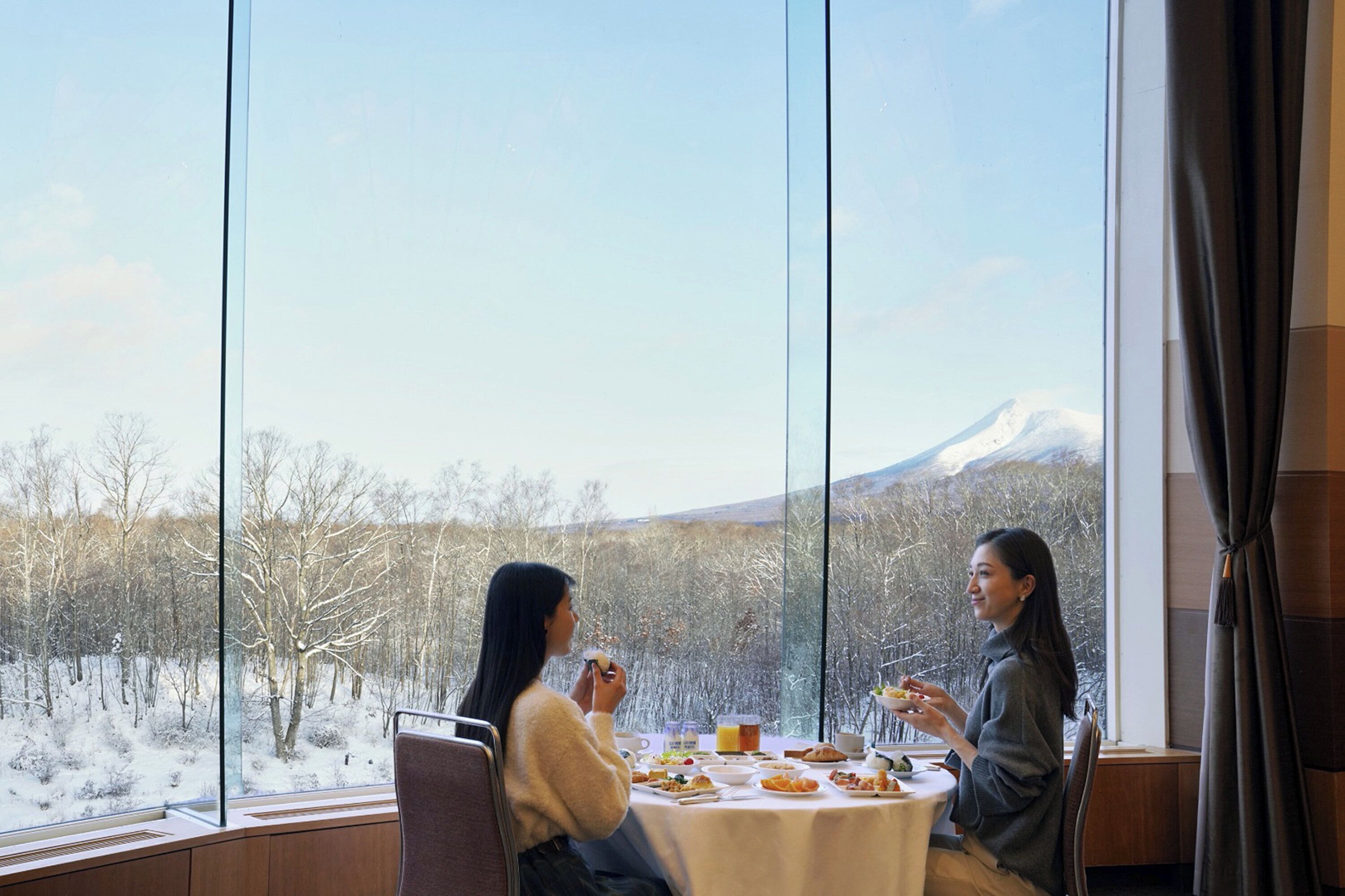 A dining area with a table set for two, large windows offering a view of a snowy landscape and a mountain.