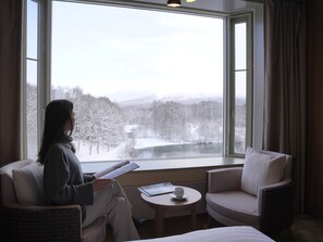 A room with a large window, a chair, a small table with a cup, and a person reading a book. - Hakodate Onuma Prince Hotel (Nanae)