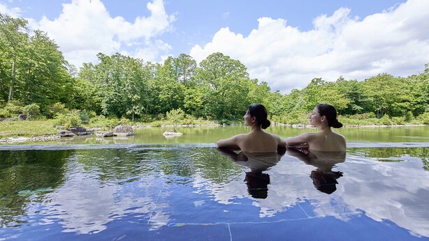 Two people in a clear, reflective pool with a natural landscape and trees in the background. - Hakodate Onuma Prince Hotel (Nanae)