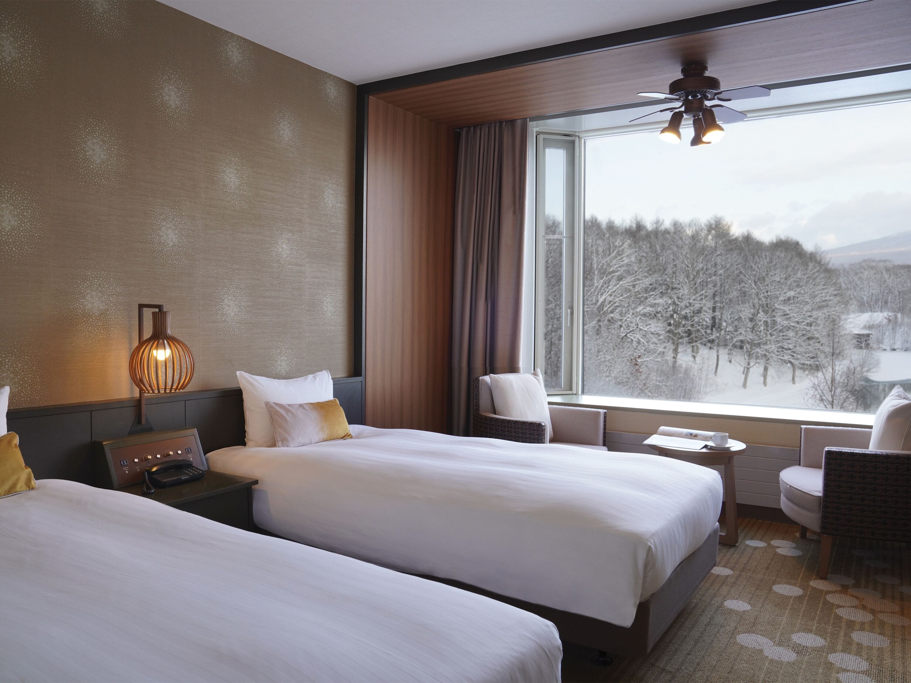 A hotel room with two beds, a desk, a chair, a lamp, and a large window with a view of a snowy landscape.