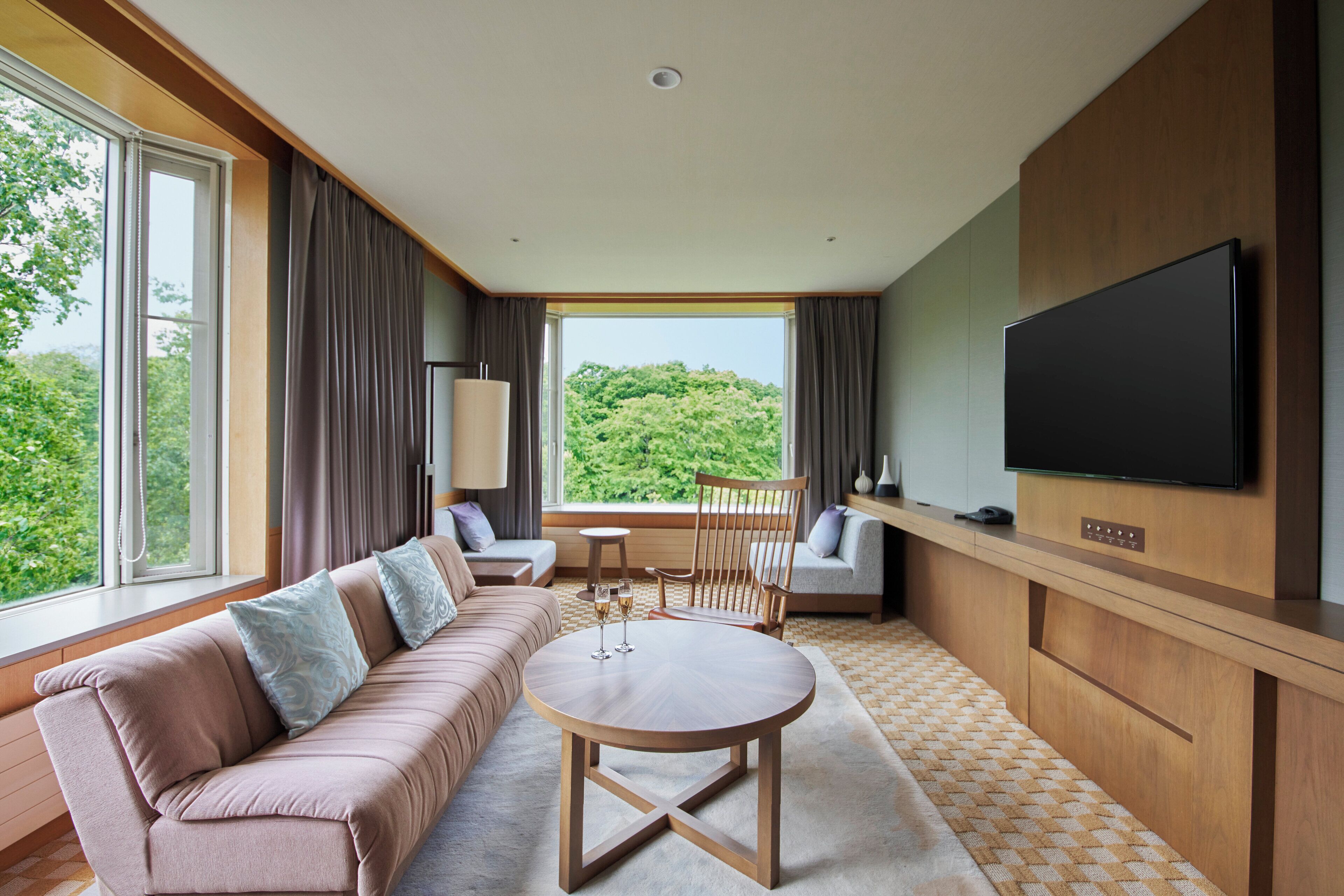 A modern hotel room with a sofa, a round table, a TV, and a view of greenery outside.