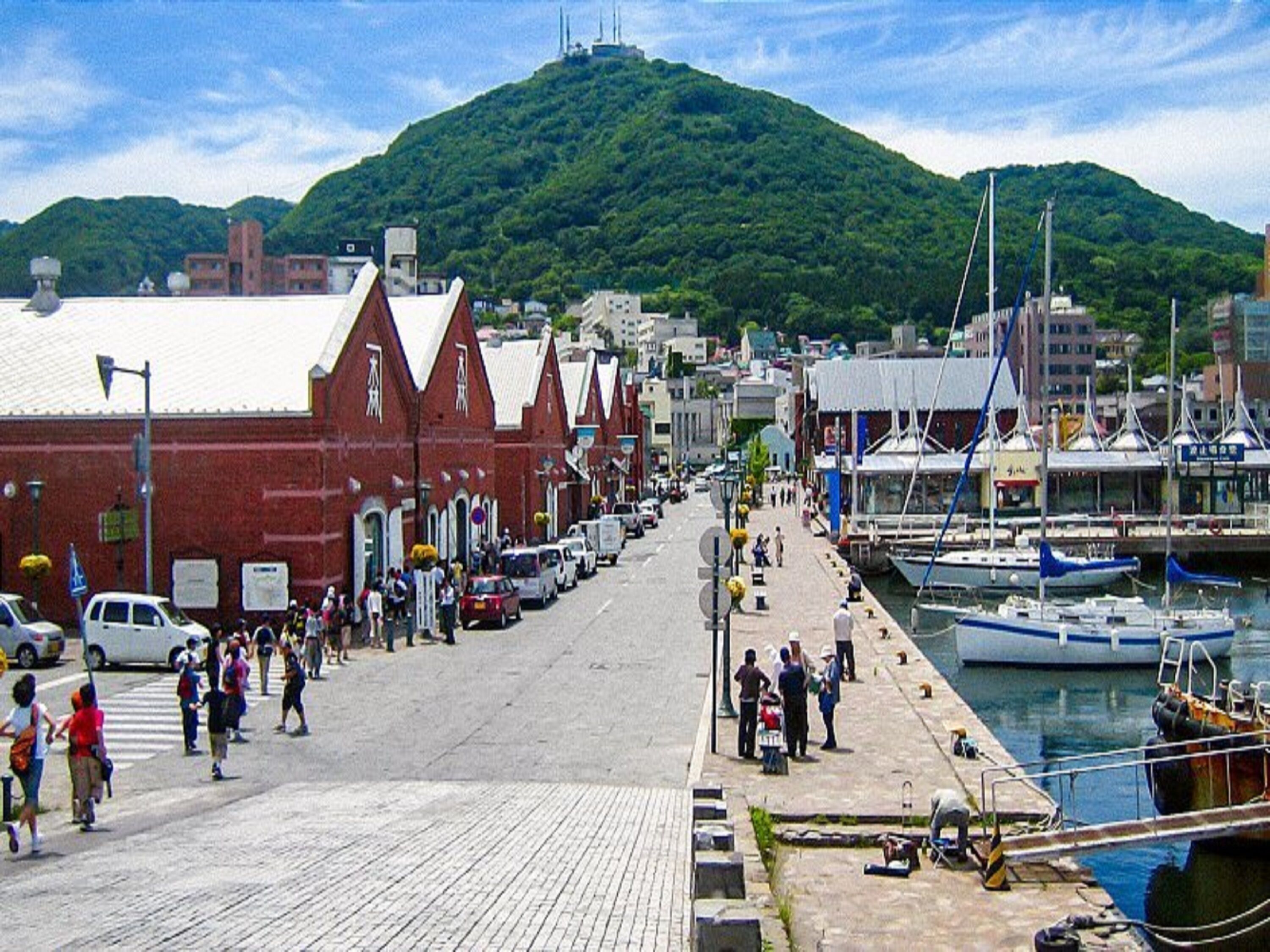 A waterfront street with buildings, a dock with boats, and a hill in the background.