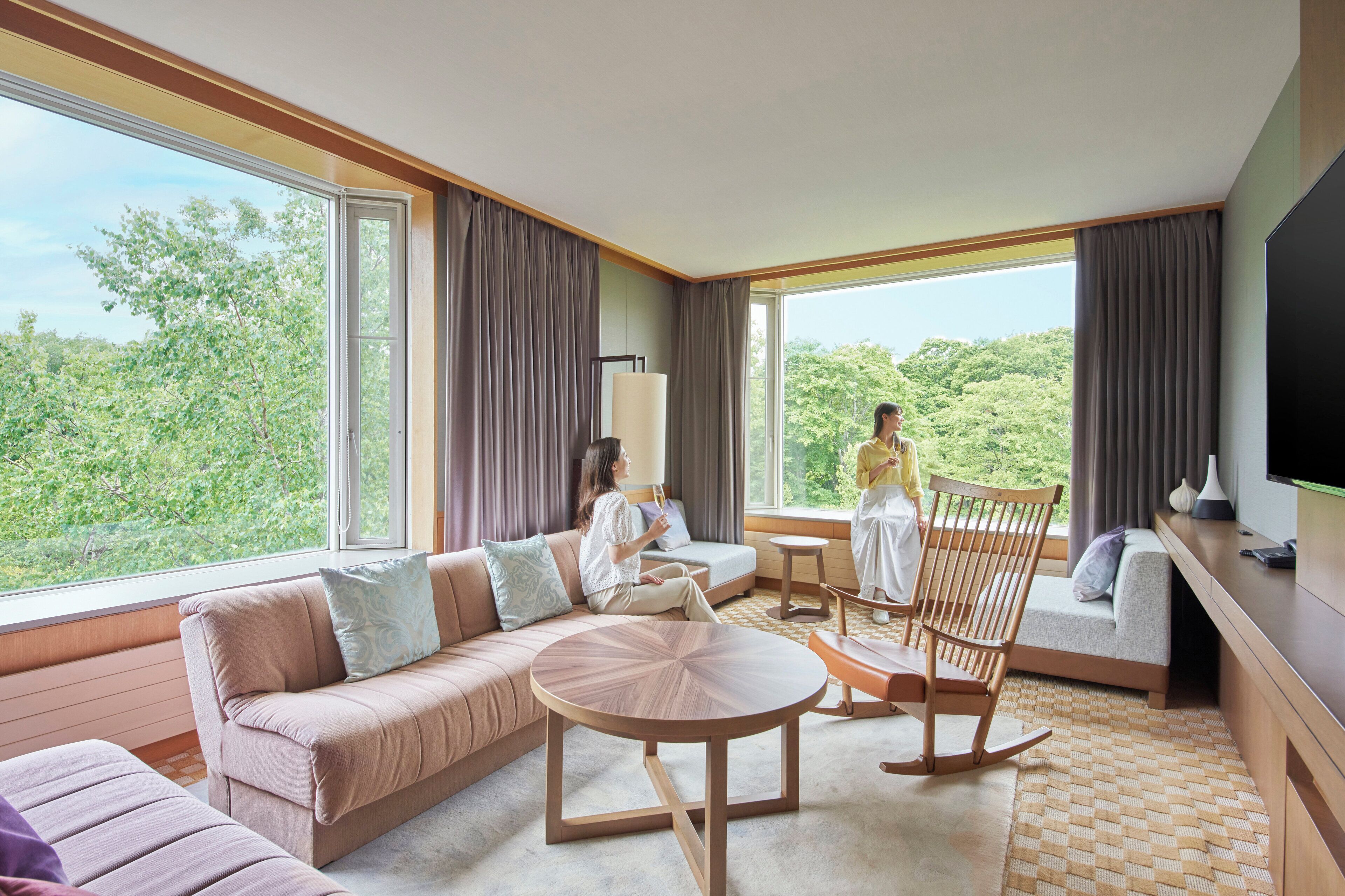 A modern hotel room with a large window, a sofa, a rocking chair, and a flat-screen TV.