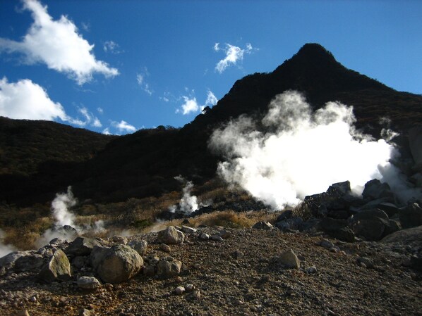 A volcanic landscape with steam rising from the ground, surrounded by rocks and sparse vegetation. - Ryuguden (Hakone)
