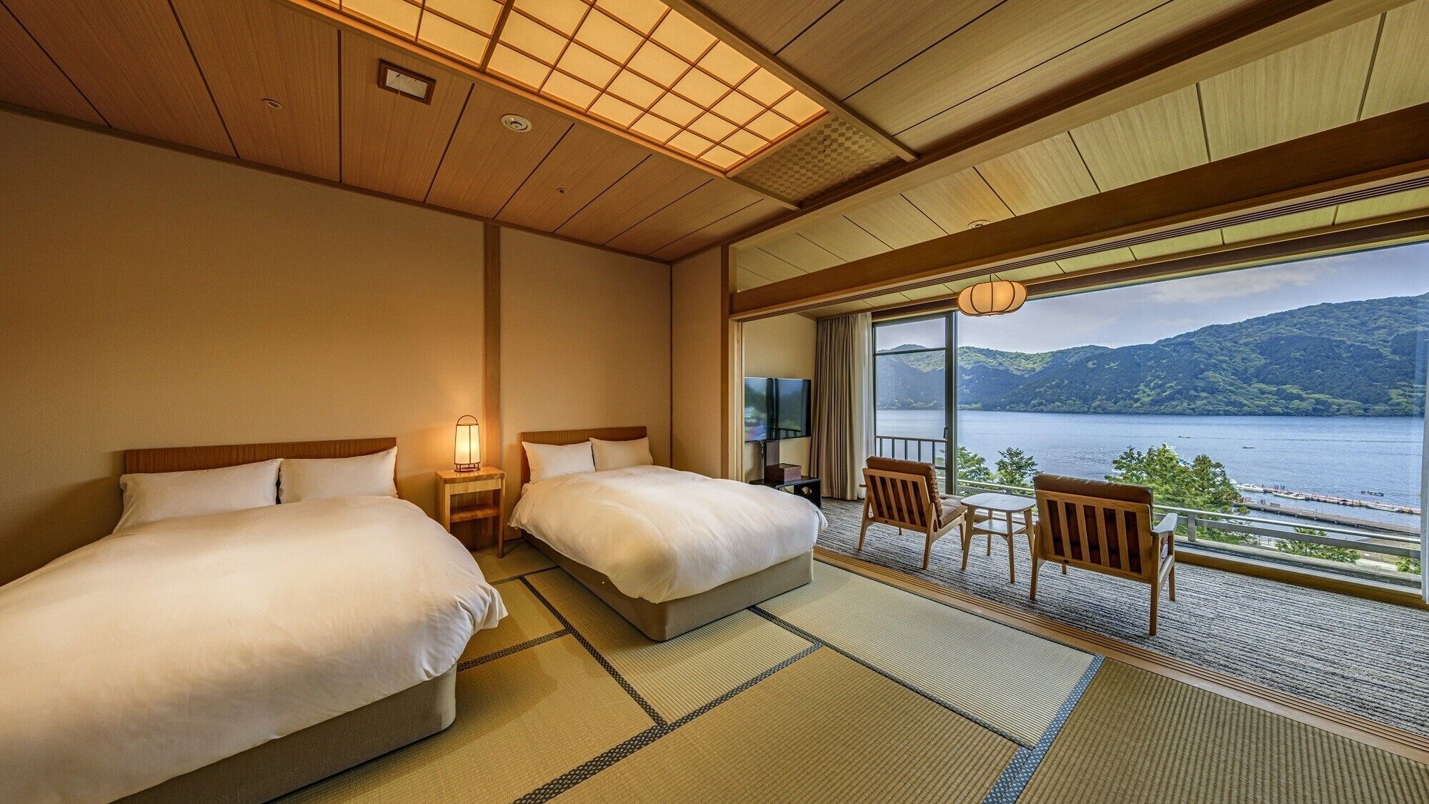 A hotel room with two beds, a TV, a balcony with a view of a lake and mountains, and traditional Japanese tatami flooring.
